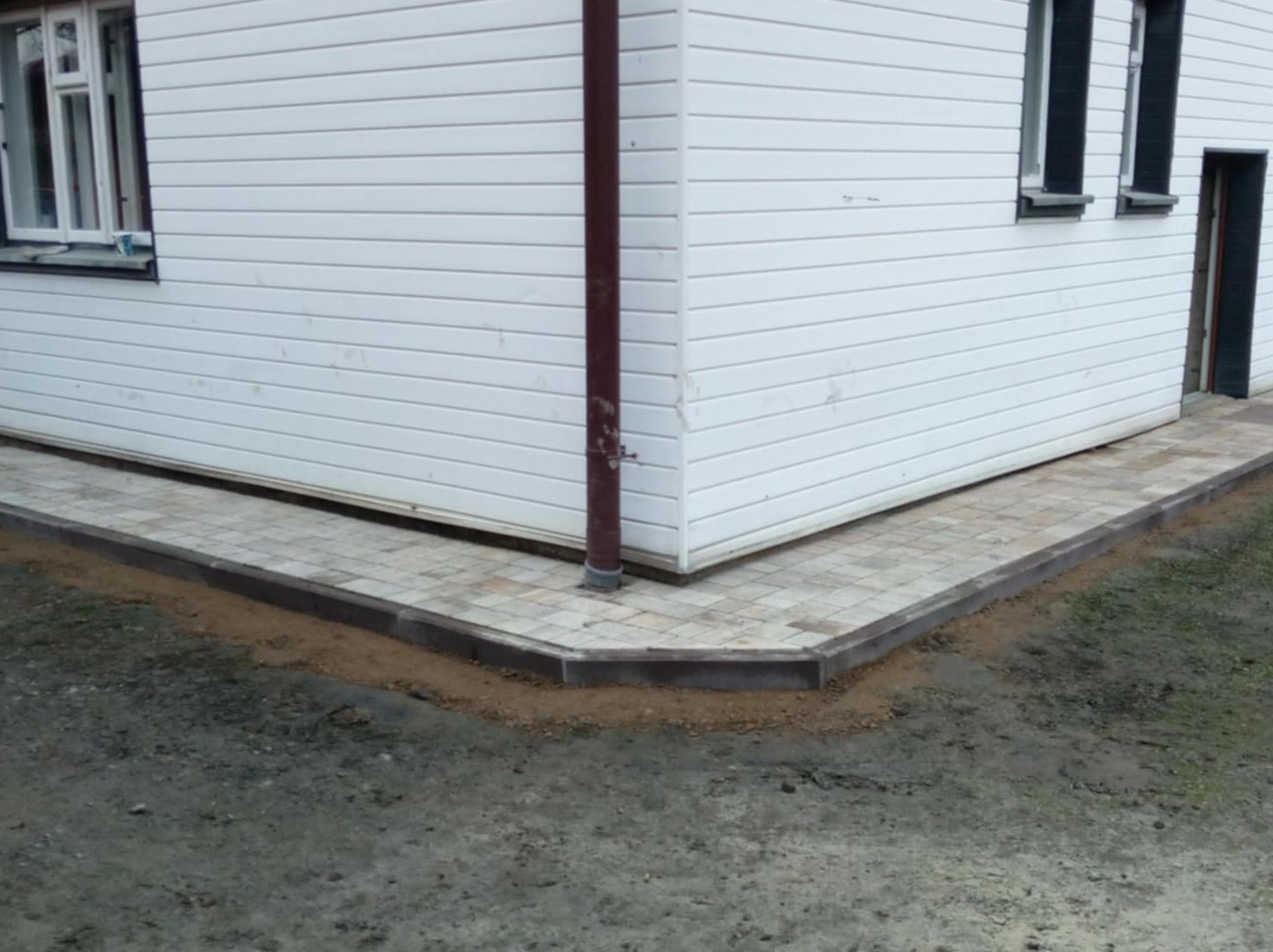 Świeżo ułożona, beżowa kostka brukowa z ciemnym obrzeżem, przylegająca do białego domu z brązową rynną, widoczne pozostałości piasku po fugowaniu.