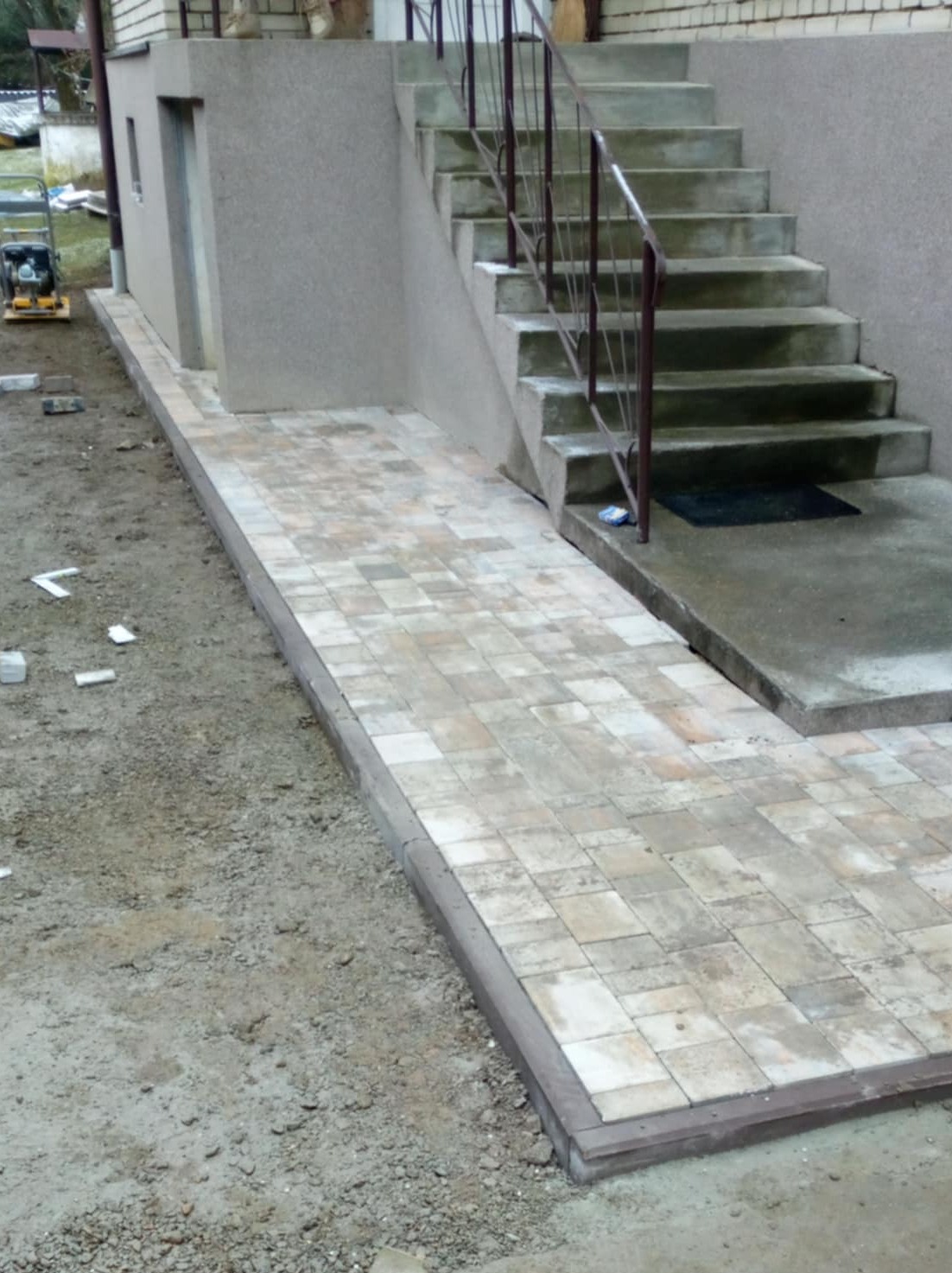 Ułożona ścieżka z kostki brukowej prowadząca do schodów wejściowych do budynku, z widoczną ubijarką w tle.