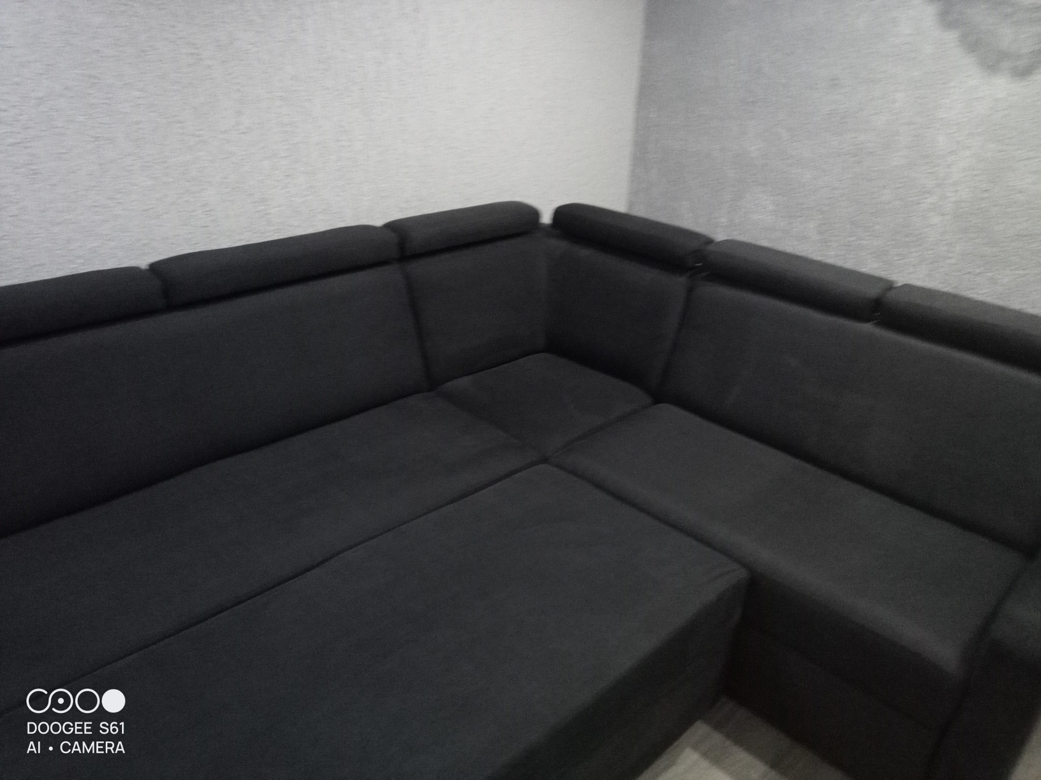 Ciemnoszara, narożna kanapa z zagłówkami w rogu pokoju, sfotografowana z niskiego kąta, widoczne lekkie niedoskonałości na ścianie.