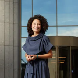 Portret uśmiechniętej kobiety w niebieskiej sukience na tle nowoczesnego biurowca z obrotowymi drzwiami i odbijającym się niebem w szklanej fasadzie.