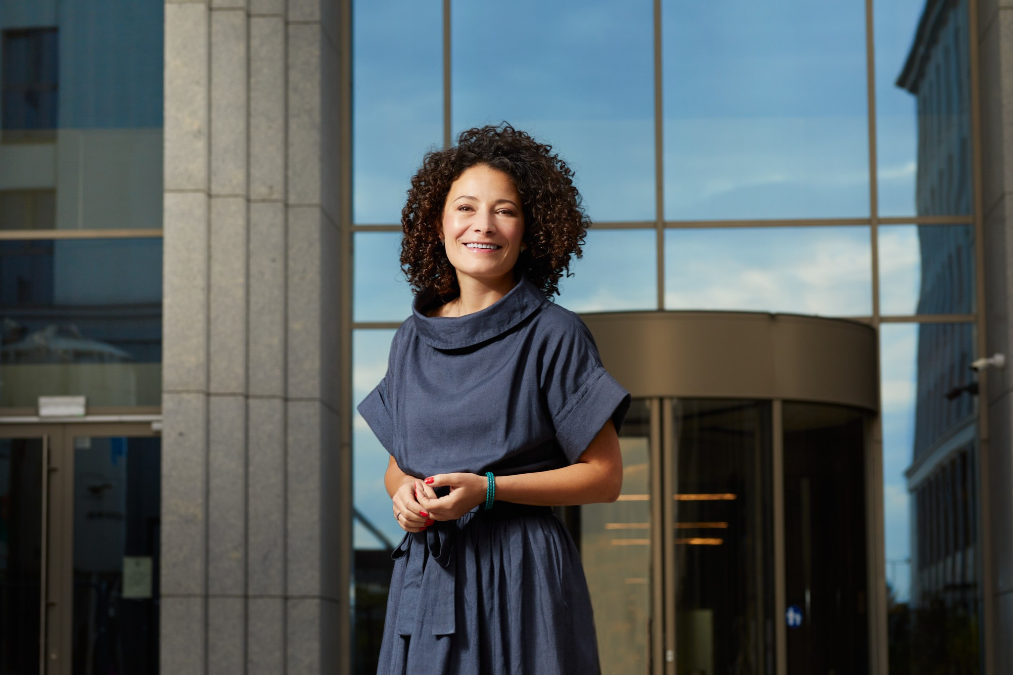 Portret uśmiechniętej kobiety w niebieskiej sukience na tle nowoczesnego biurowca z obrotowymi drzwiami i odbijającym się niebem w szklanej fasadzie.