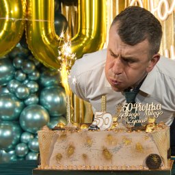MAR-MAL Mariusz Piórkowski - Mężczyzna w Głownie zdmuchuje płomień z tortu urodzinowego z okazji 50-tych urodzin, w tle balony w kolorze złota i turkusu oraz złota kurtyna.
