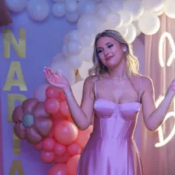 Kobieta w różowej sukni na tle dekoracji urodzinowych: balony, kwiaty, neon z napisem 'Happy Birthday'. Uśmiechnięta, z rozłożonymi rękami. Sesja urodzinowa w studio.