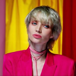 MAR-MAL Mariusz Piórkowski - Portret kobiety w różowym żakiecie na tle kolorowych zasłon. Krótkie blond włosy, wyrazisty makijaż oczu i ust. Profesjonalna sesja w studio.