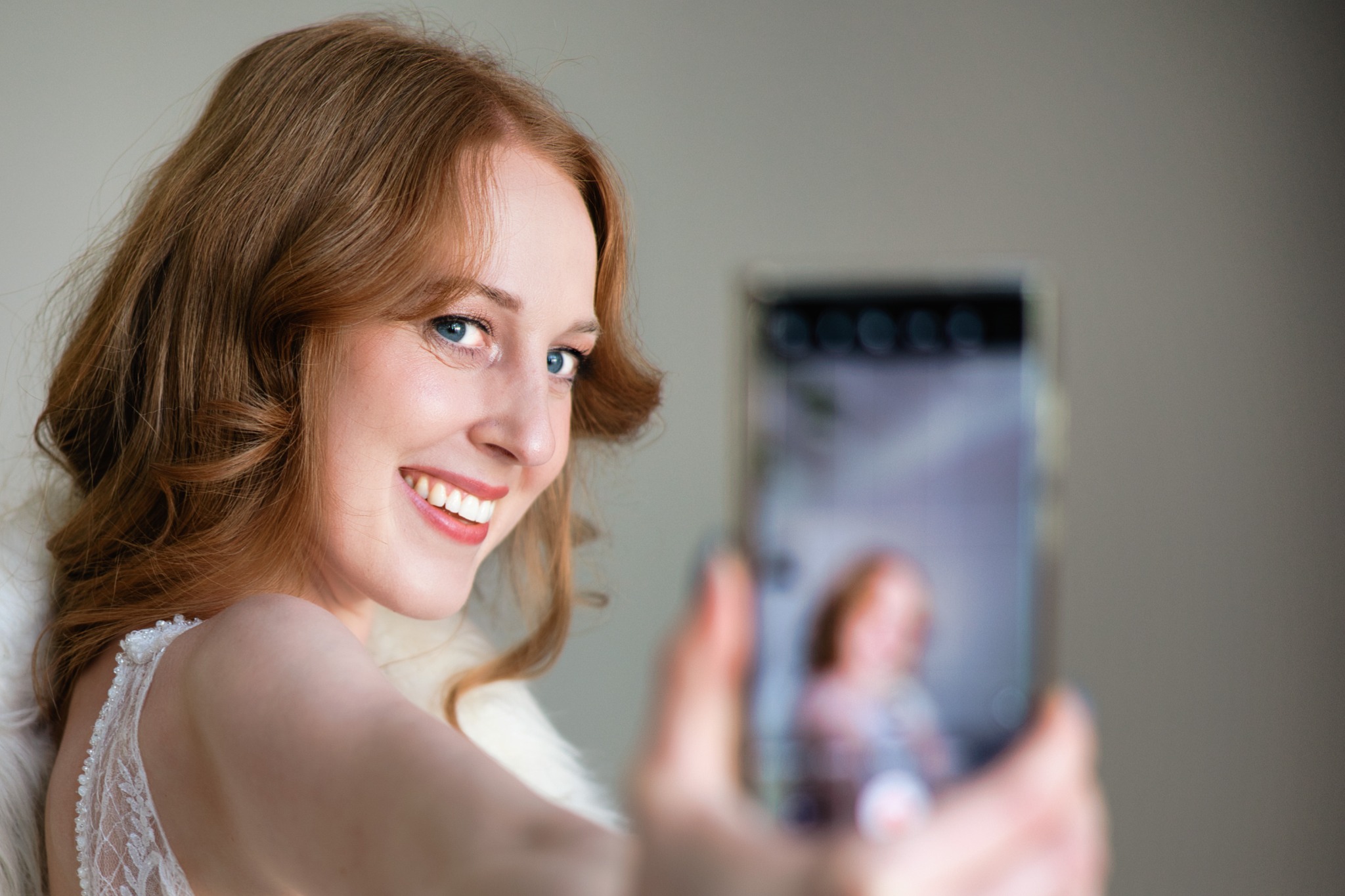 Uśmiechnięta rudowłosa kobieta w białej koronkowej sukience pozuje do selfie, trzymając telefon komórkowy, na którego ekranie widoczny jest jej rozmazany wizerunek.