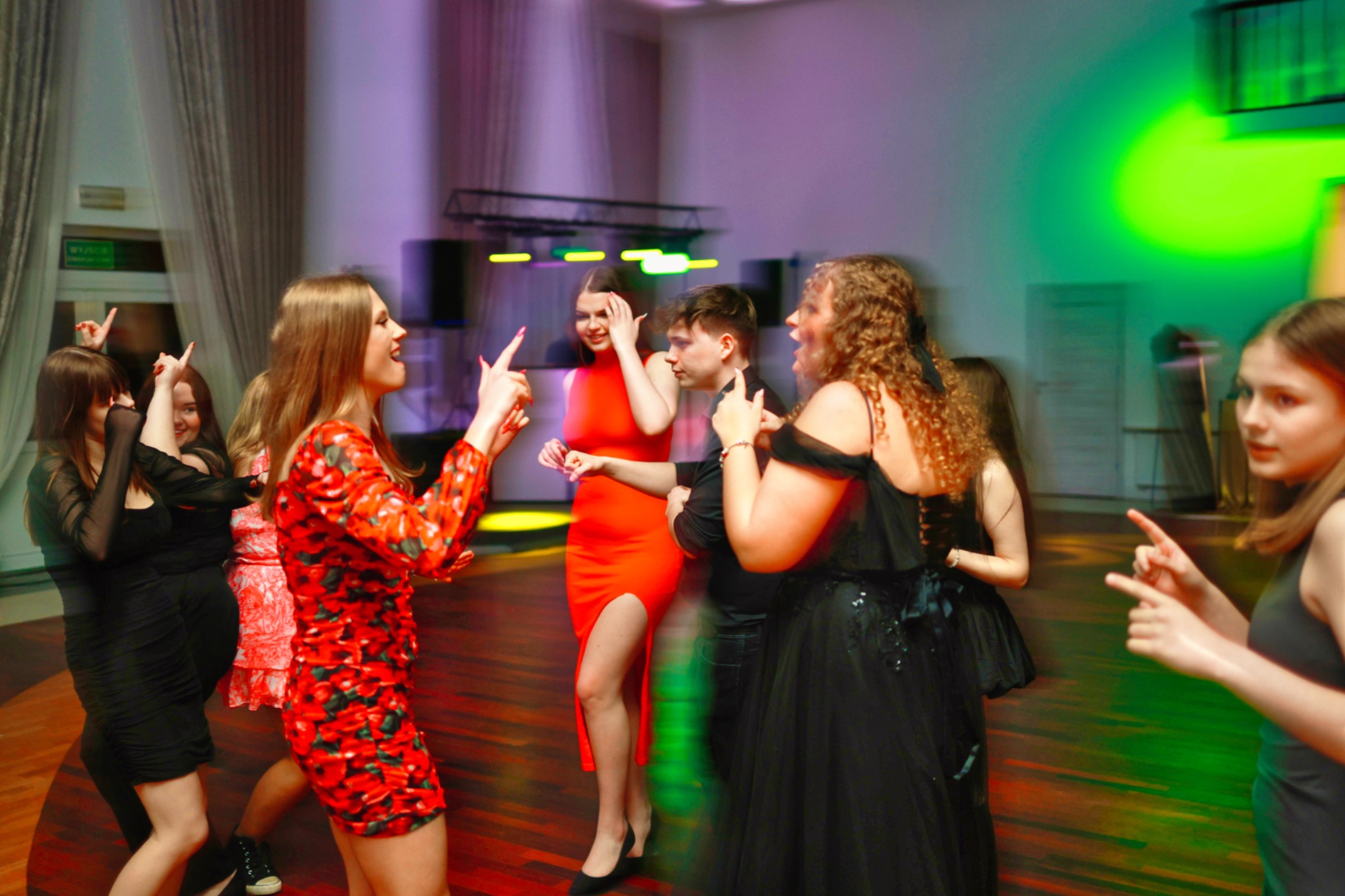 Roztańczona młodzież w sukienkach na parkiecie, oświetlona kolorowymi światłami, w tle okna z zasłonami i sprzęt nagłośnieniowy.