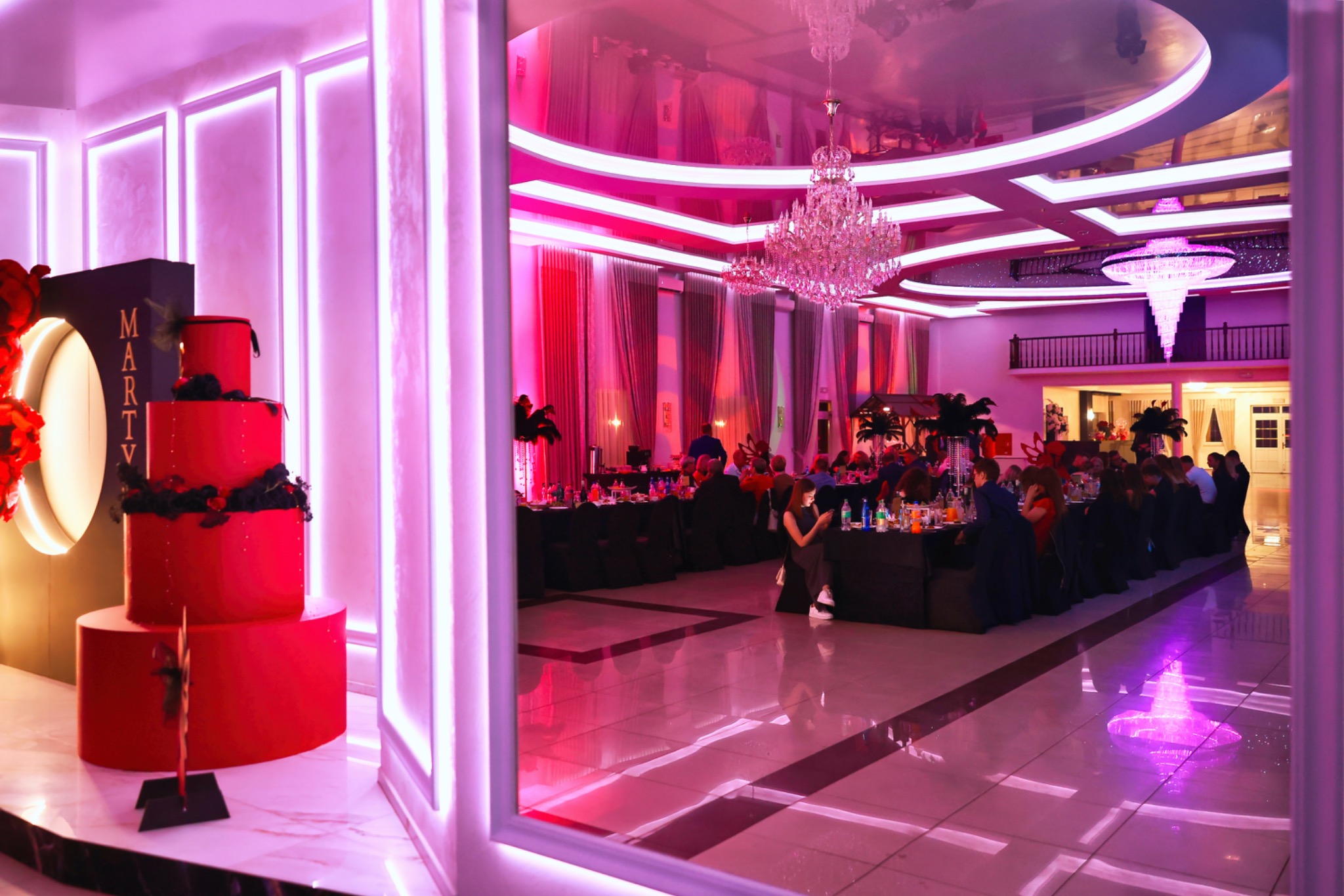 Elegancka sala bankietowa z czerwonym tortem 'Marty', kryształowe żyrandole, oświetlenie LED i goście przy stołach. Ujęcie zza ozdobnej ramy.