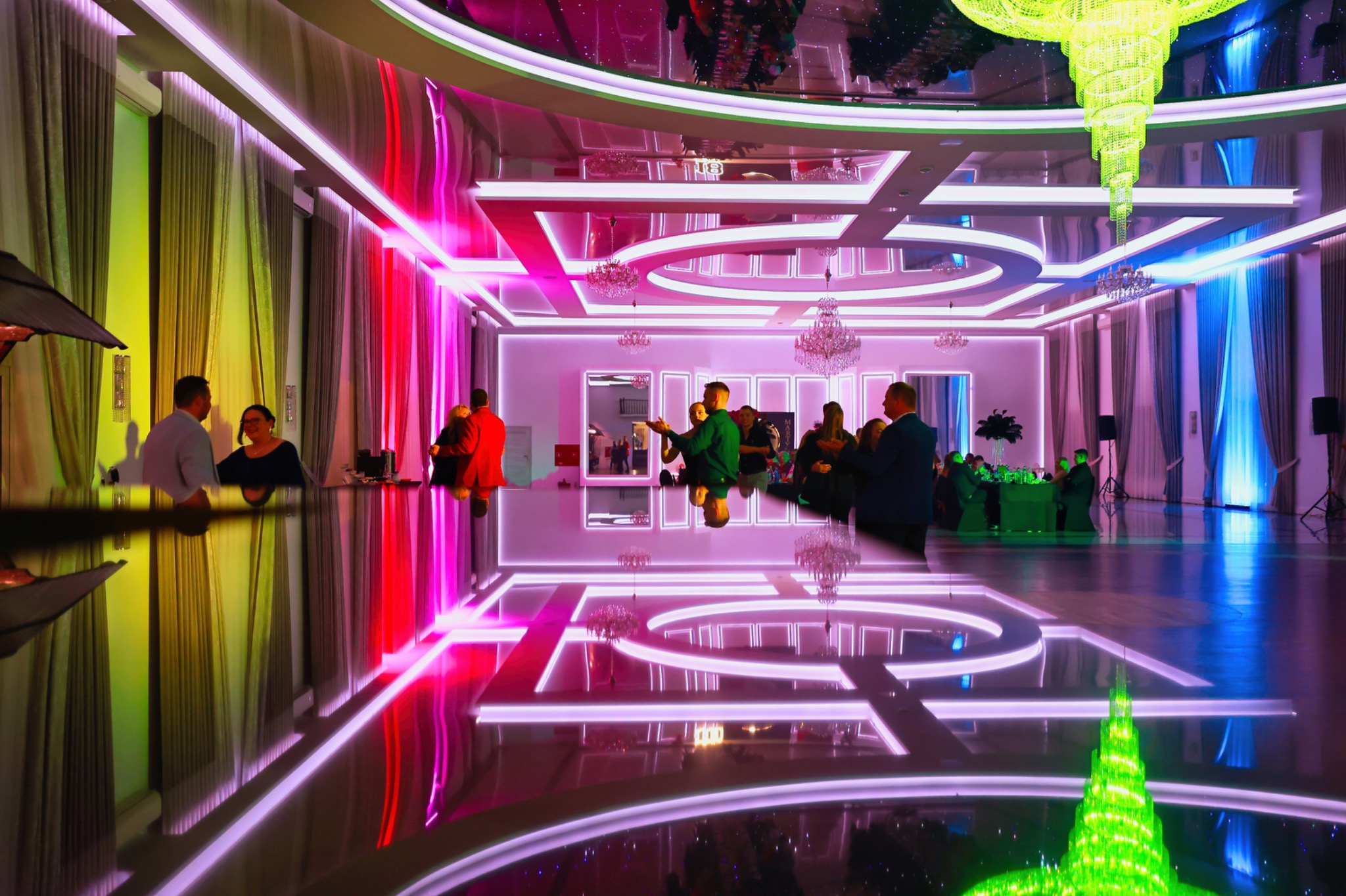 Elegancka sala bankietowa z lustrzanym sufitem, oświetlona kolorowymi światłami LED i kryształowymi żyrandolami, przygotowana na imprezę. Na sali znajdują się goście.