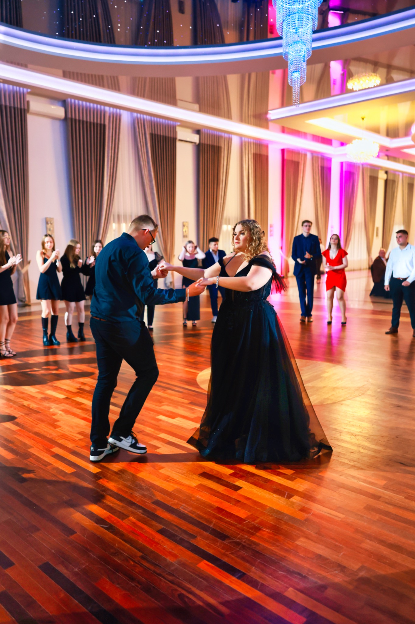 Para tańczy na parkiecie w sali balowej z kryształowym żyrandolem. W tle goście. Kobieta w czarnej sukni, mężczyzna w ciemnej koszuli i sportowych butach. Parkiet z desek.