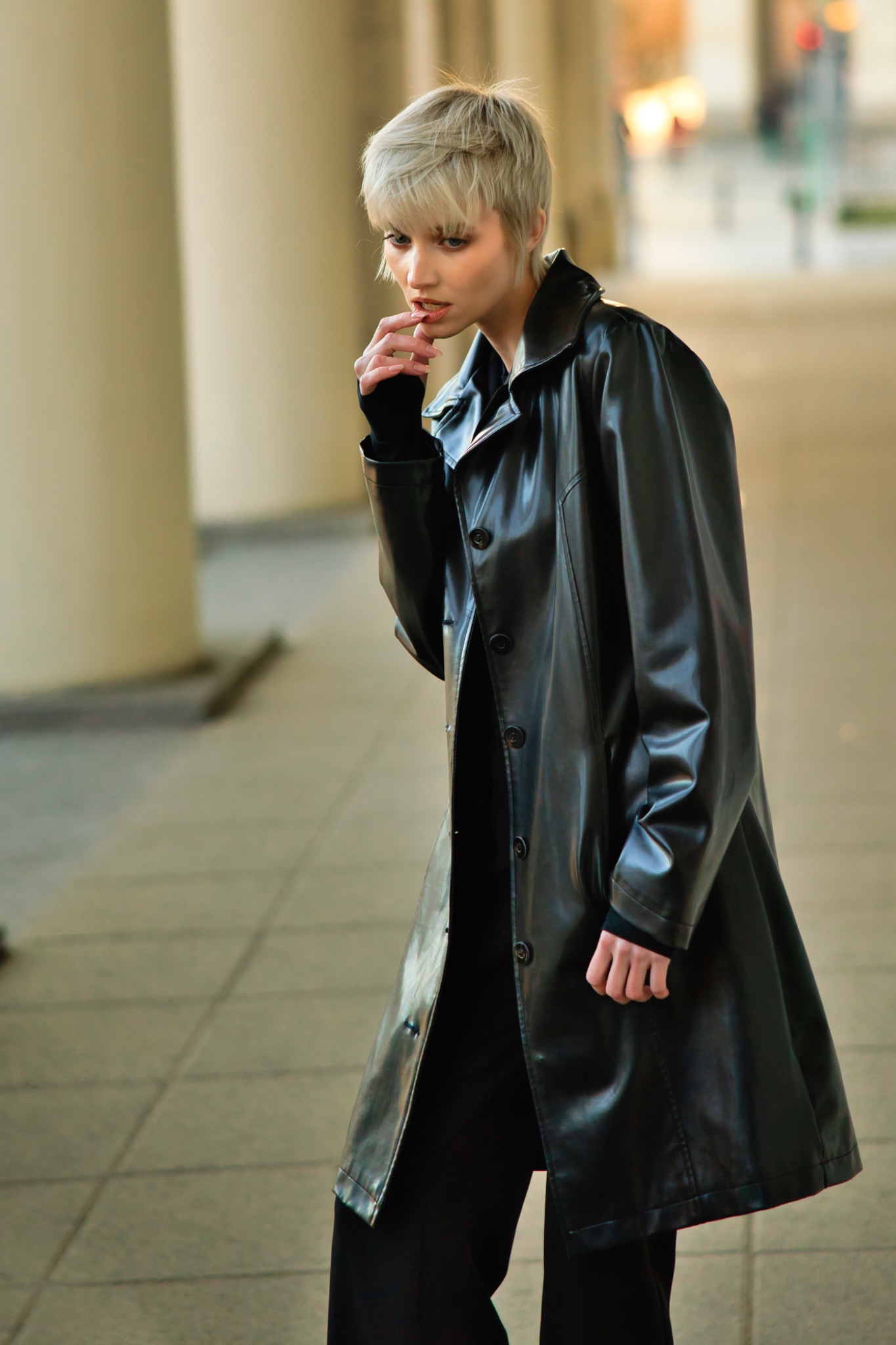 Portret kobiety w czarnym, lakierowanym płaszczu na tle kolumn. Krótkie blond włosy, zamyślony wyraz twarzy. Sesja w miejskiej przestrzeni.