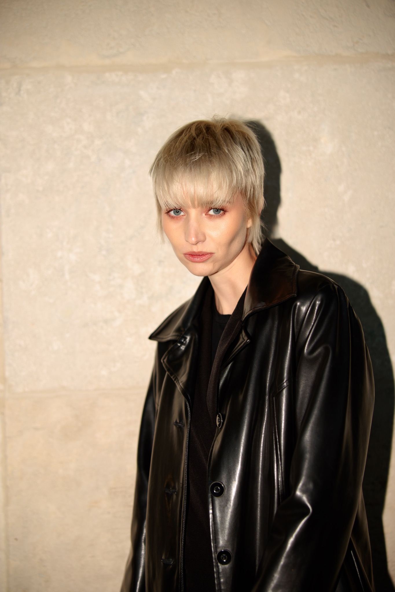 Portret kobiety z krótkimi blond włosami w czarnej, skórzanej kurtce na tle jasnej ściany. Uwagę przykuwają jej niebieskie oczy i delikatny makijaż.