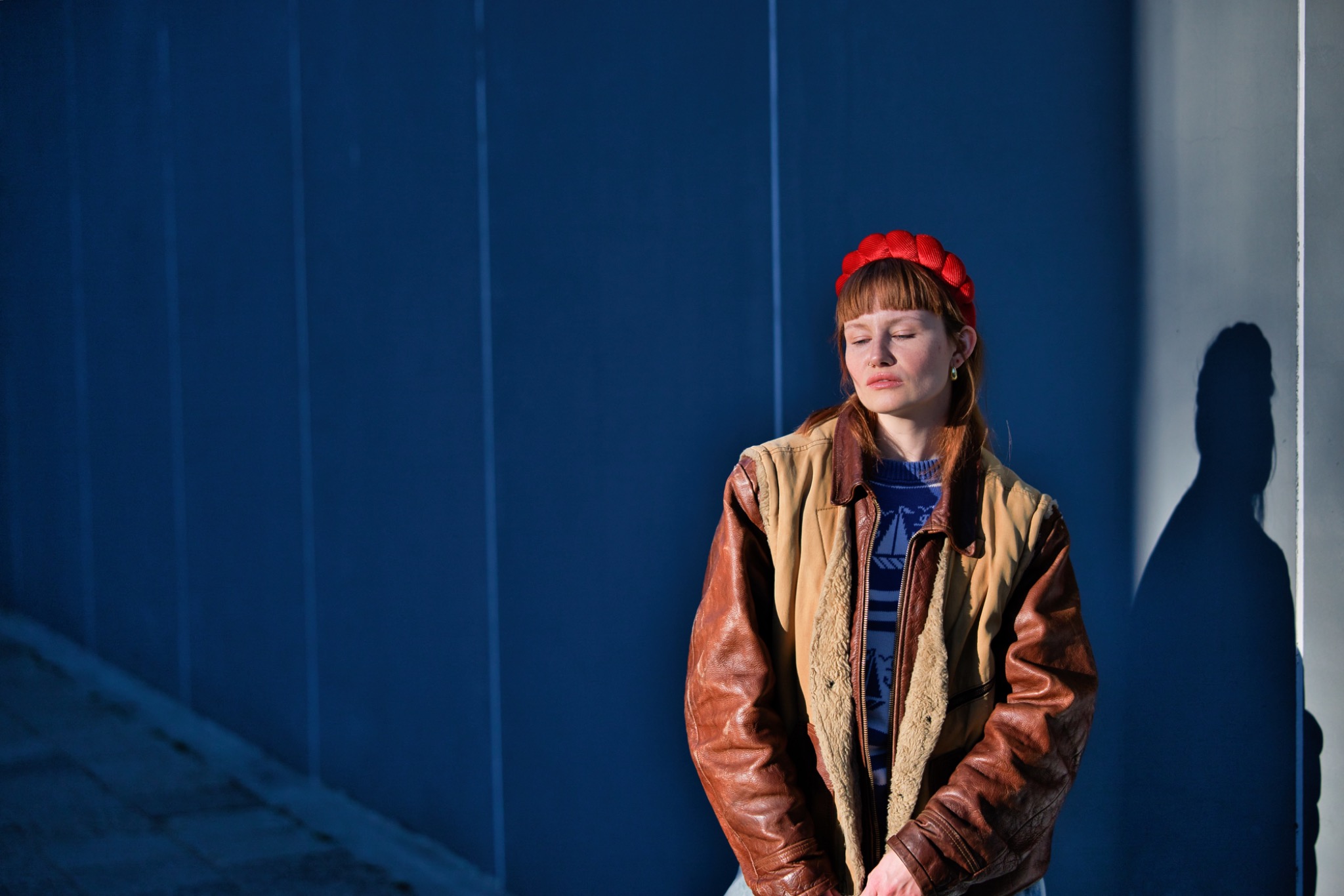 Portret kobiety z rudymi włosami i czerwoną opaską na tle niebieskiej ściany w słoneczny dzień. Na ścianie widoczny cień osoby.