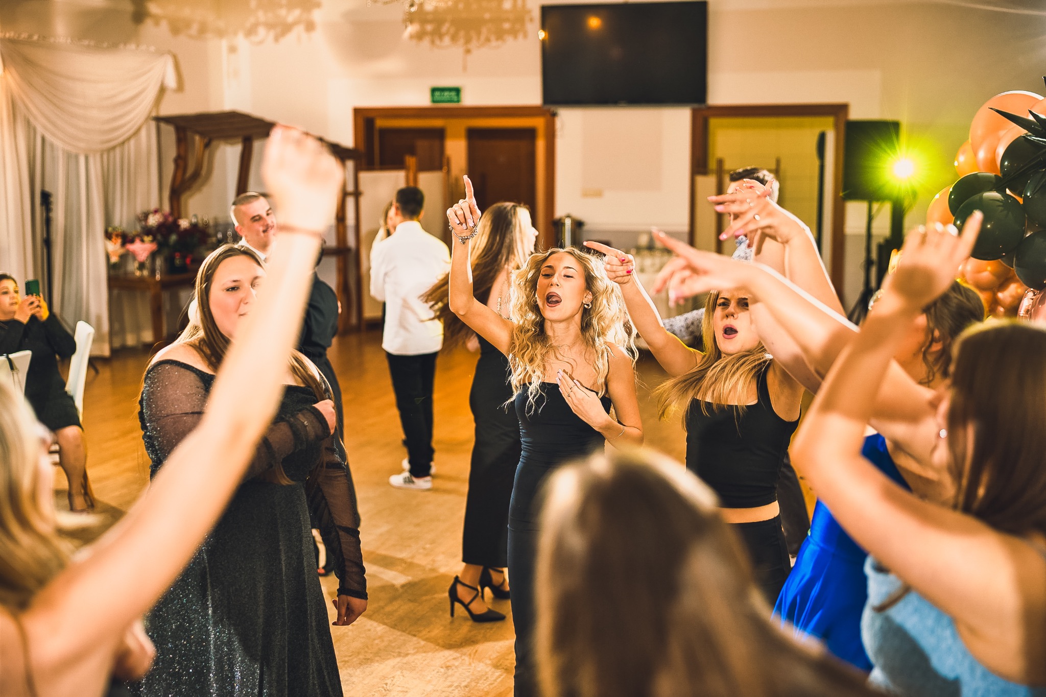 Roztańczona grupa kobiet w sukienkach na parkiecie, w tle dekoracje i światła. Uczestnicy imprezy w ruchu, z uniesionymi rękami i ekspresyjnymi minami.
