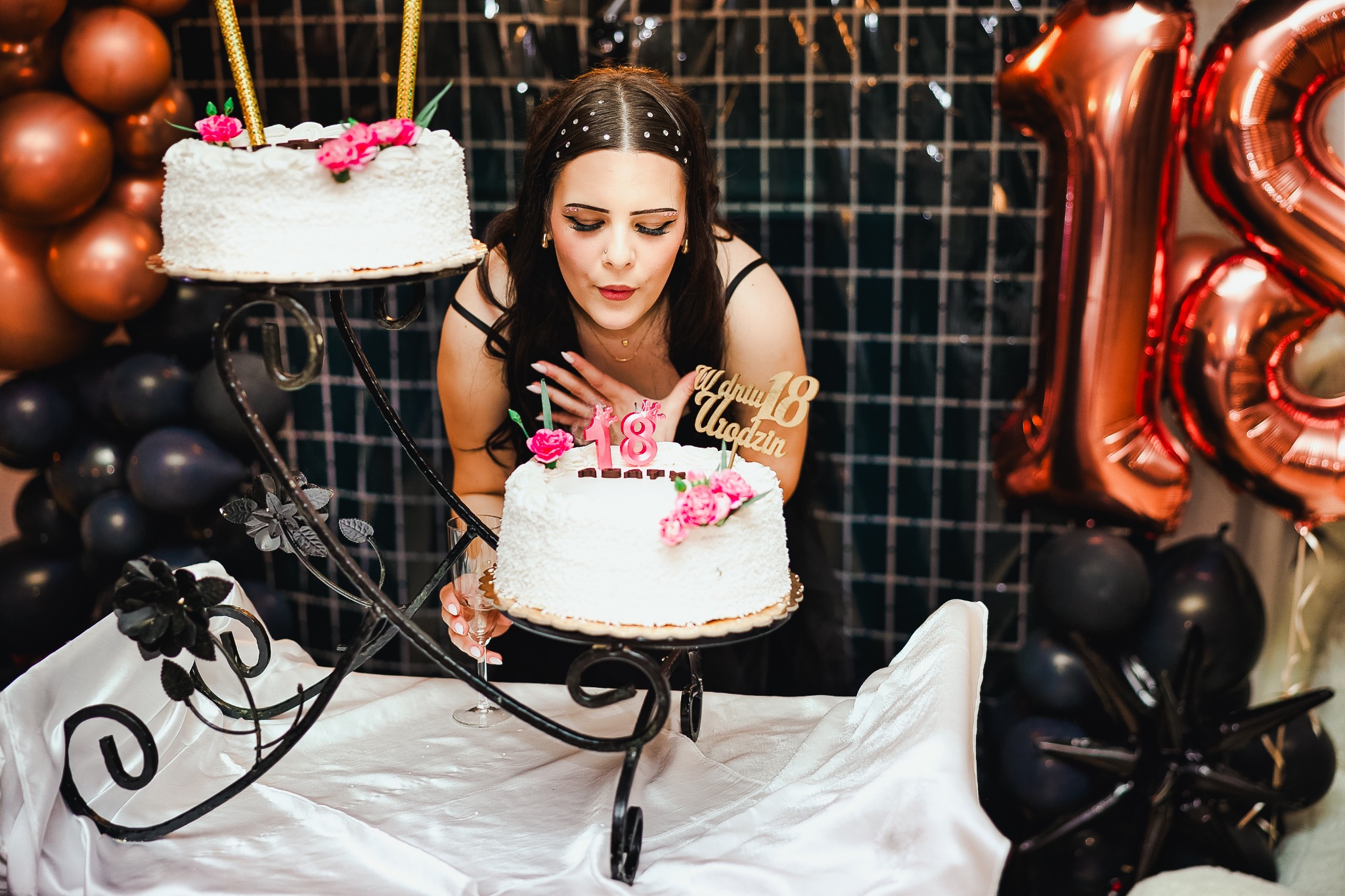 Uśmiechnięta młoda kobieta dmucha świeczki na torcie urodzinowym podczas uroczystości osiemnastych urodzin, z dekoracjami i balonami w tle.