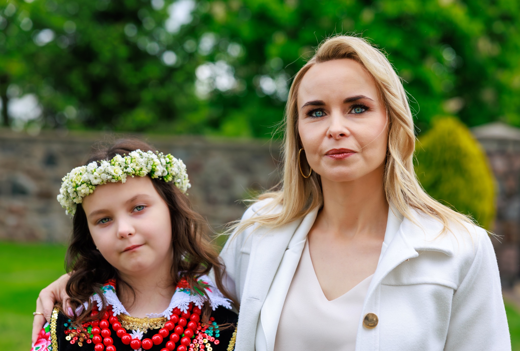 Portret kobiety i dziewczynki w stroju ludowym z wiankiem na głowie, na tle zieleni. Jasne, naturalne światło, neutralny wyraz twarzy, skupienie na detalach stroju.