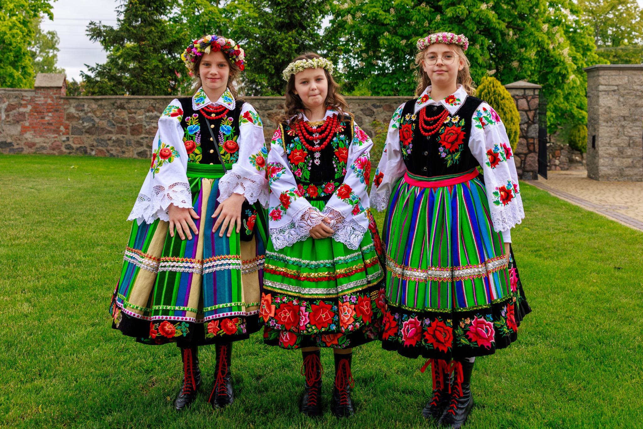 Trzy dziewczynki w barwnych, tradycyjnych strojach ludowych z kwiatowymi wiankami na głowach, stoją na trawie przed kamiennym murem i zielenią drzew.