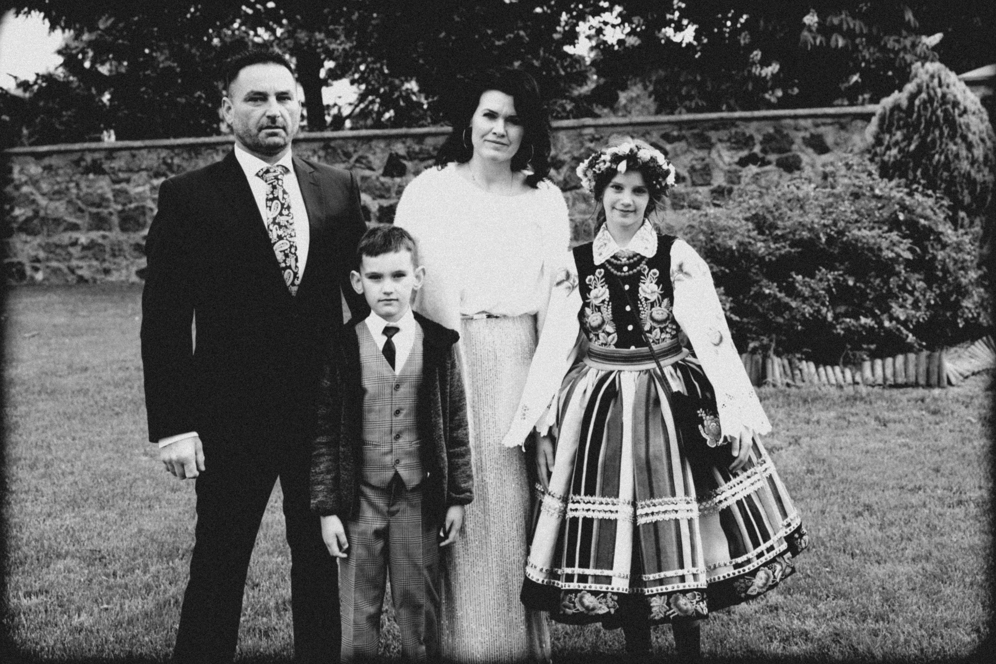 Czarno-białe zdjęcie rodzinne: mężczyzna w garniturze, kobieta i dwoje dzieci, dziewczynka w stroju ludowym, na tle kamiennego muru i zieleni.