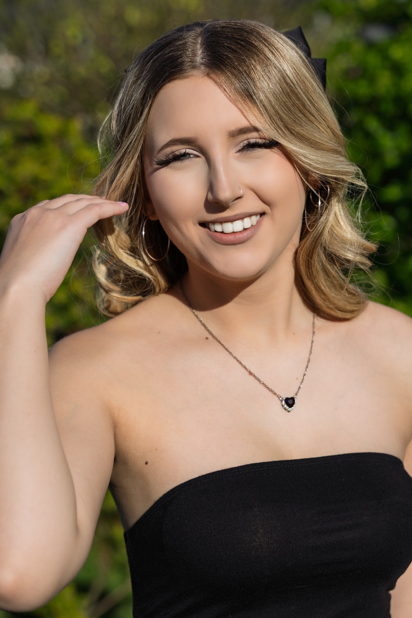 Uśmiechnięta blondynka w czarnej sukience bez ramiączek, z kolczykami kołami i naszyjnikiem w kształcie serca, pozuje na tle zieleni.