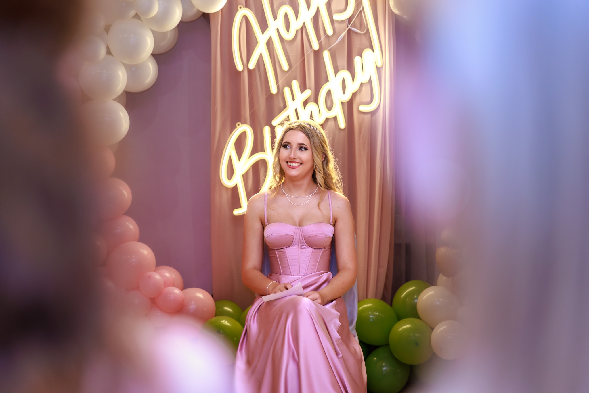 Uśmiechnięta blondynka w różowej sukni siedzi na tle dekoracji urodzinowej z balonów i neonu 'Happy Birthday'.