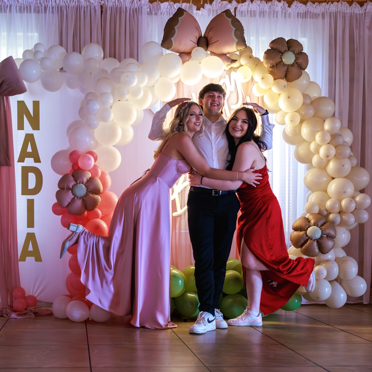 Uśmiechnięci młodzi ludzie w objęciach na tle dekoracji z balonów i kwiatów w odcieniach różu i beżu. Dziewczyny w sukienkach wieczorowych, chłopak w białej koszuli.