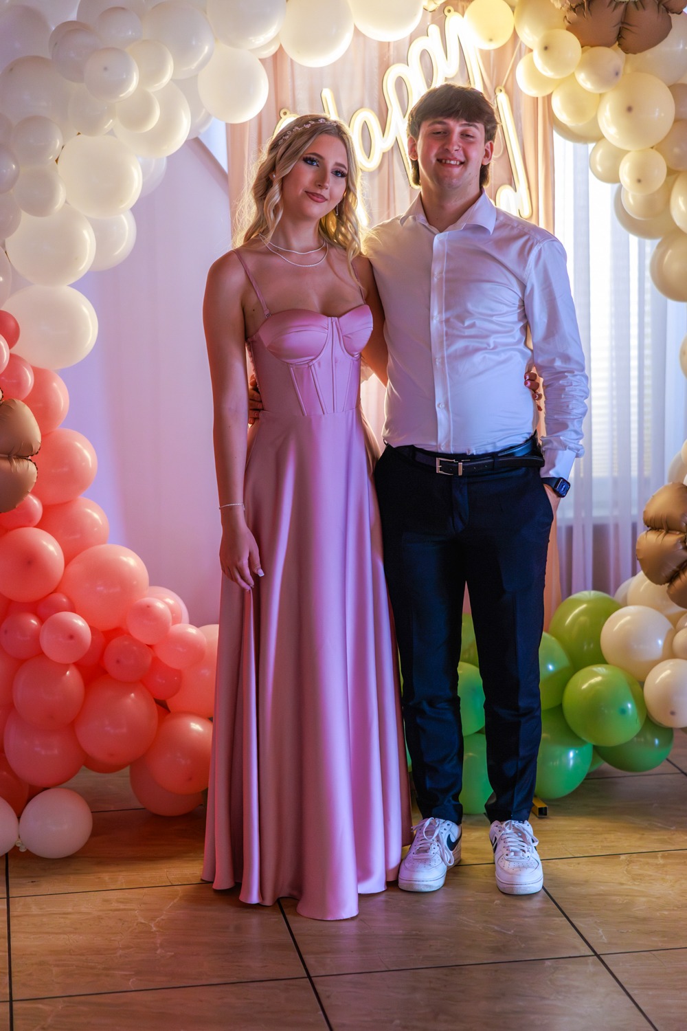 Para ubrana elegancko, ona w długiej, różowej sukni, on w białej koszuli i czarnych spodniach, pozuje na tle dekoracji z balonów.