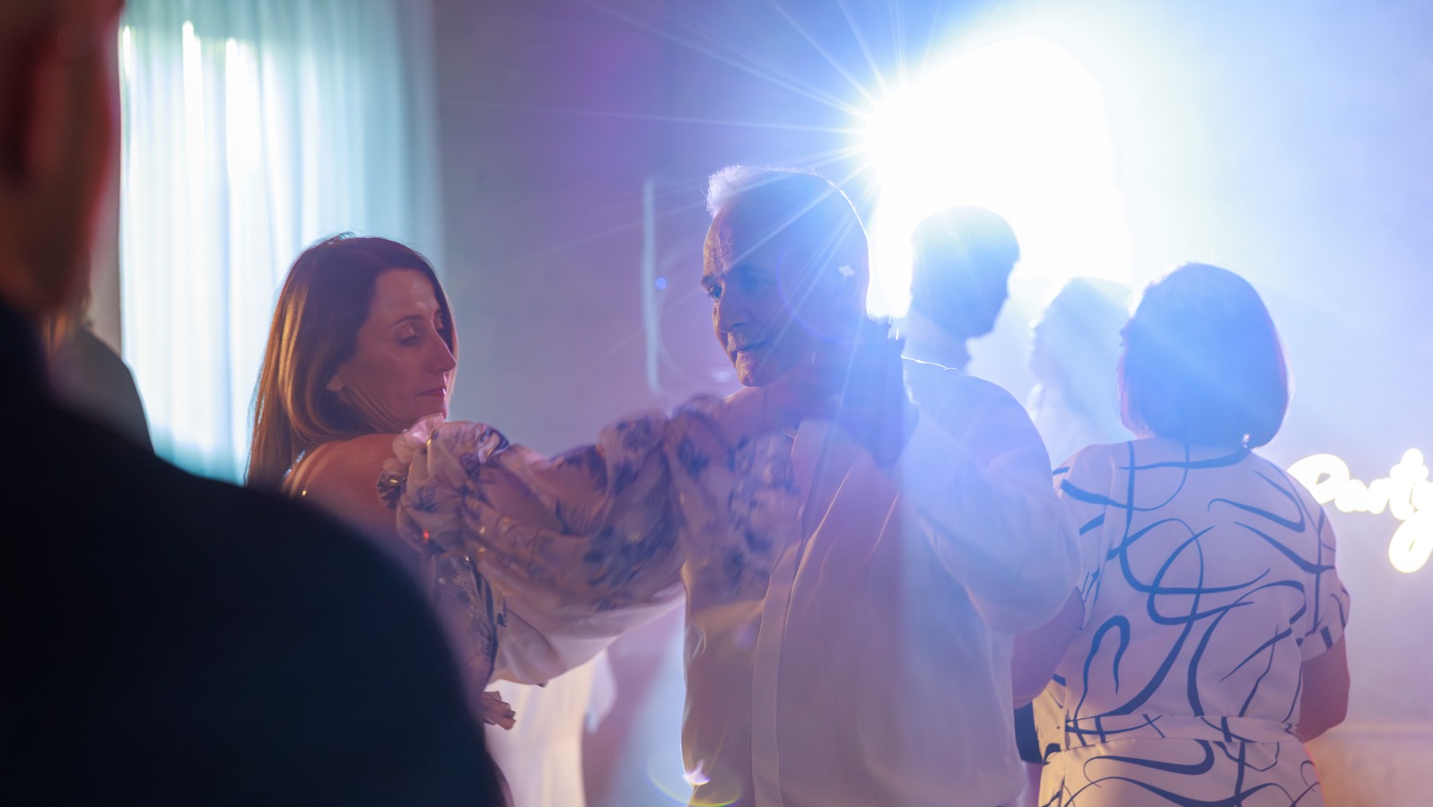 Para tańczy na parkiecie podczas imprezy, oświetlona światłem reflektorów i neonowym napisem 'Party' w tle. Widoczni inni goście.