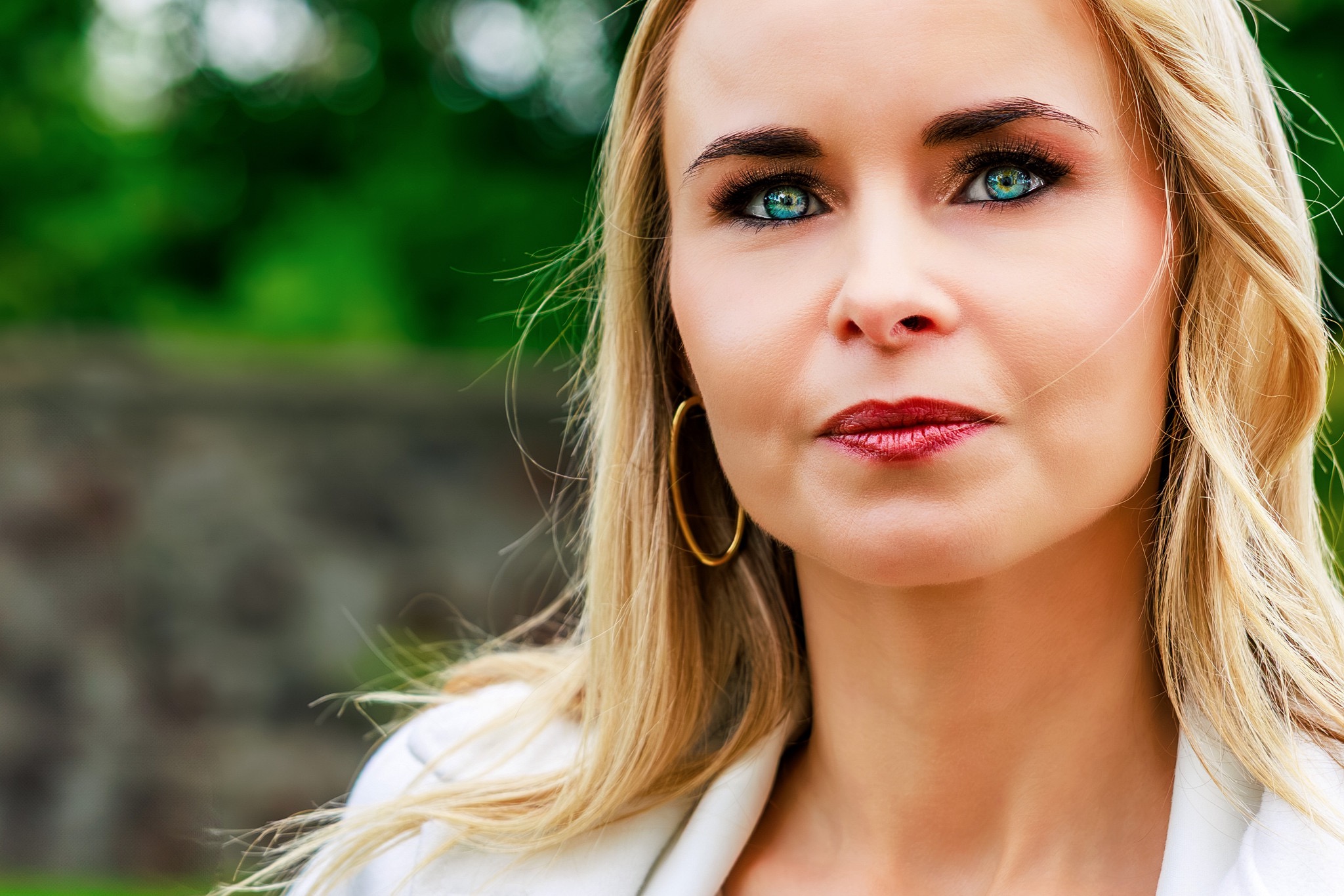 Portret kobiety o blond włosach i intensywnym makijażu oczu, z zielonym tłem w tle. Widoczne złote kolczyki koła i biała marynarka.