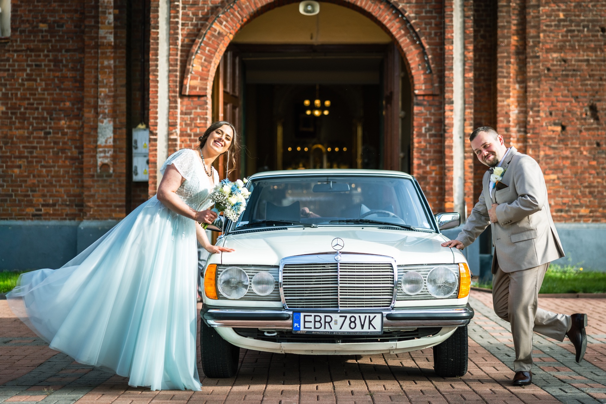 Uśmiechnięta para młoda w jasnych strojach pozuje przy białym Mercedesie przed ceglanym kościołem z otwartymi drzwiami.