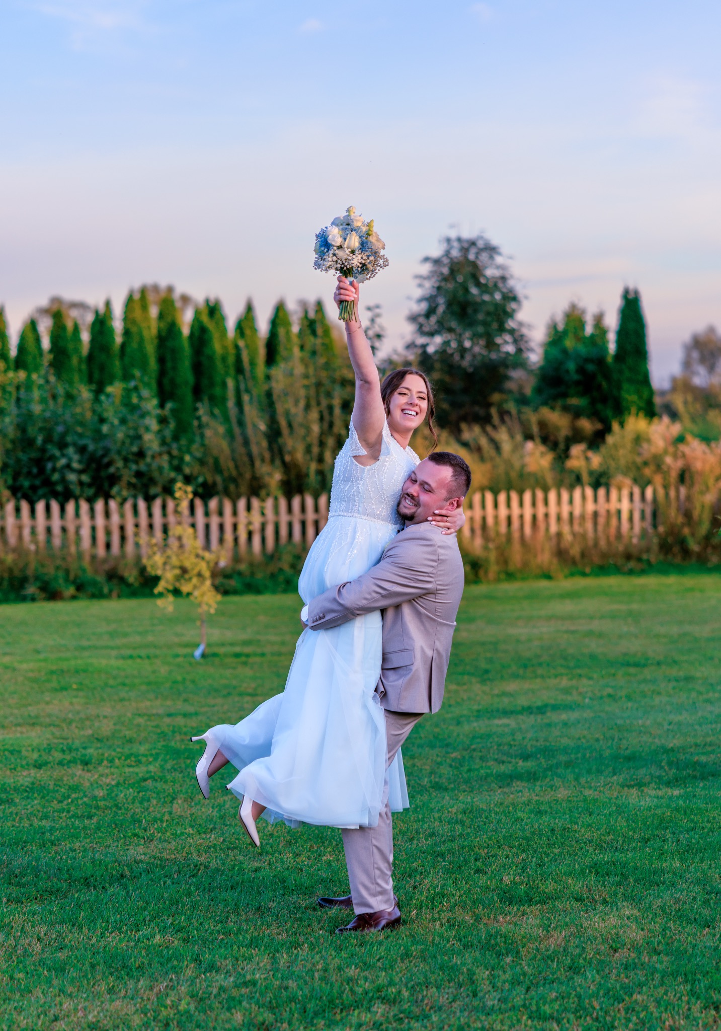 Uśmiechnięta panna młoda w białej sukni, trzymana w górze przez pana młodego w beżowym garniturze, na tle zielonego trawnika i drzew podczas sesji ślubnej.