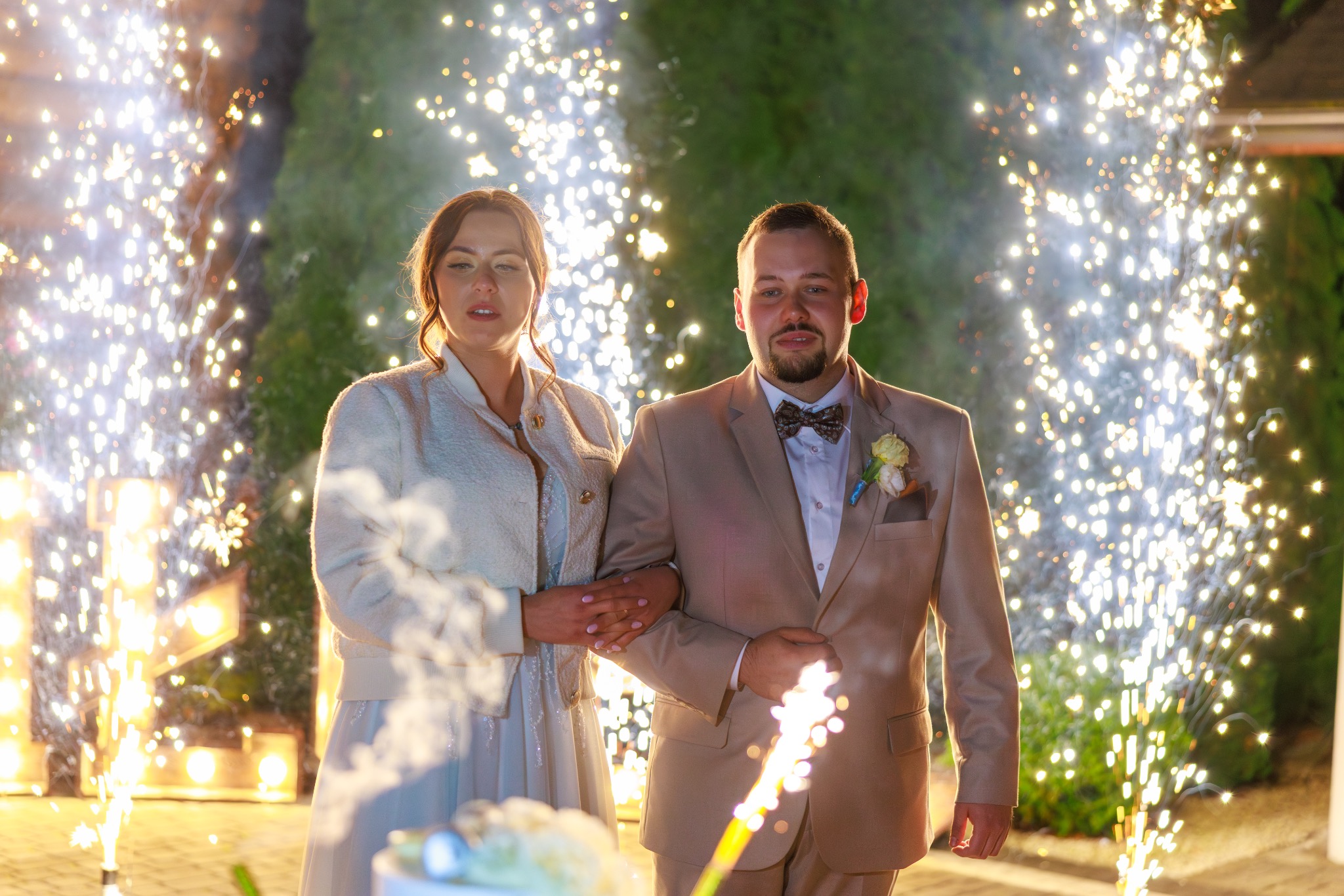Para młoda w beżowych strojach, otoczona fontannami iskier na tle zieleni. Ujęcie z dołu, podkreślające uroczysty charakter wydarzenia.