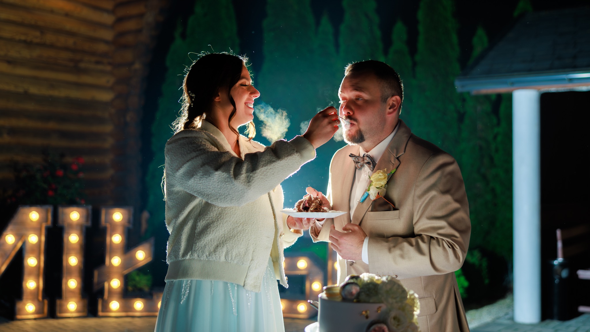 Uśmiechnięta panna młoda karmi tortem pana młodego na tle dekoracji świetlnych i zieleni. Romantyczna scena z wesela, uchwycona w ciepłym oświetleniu.