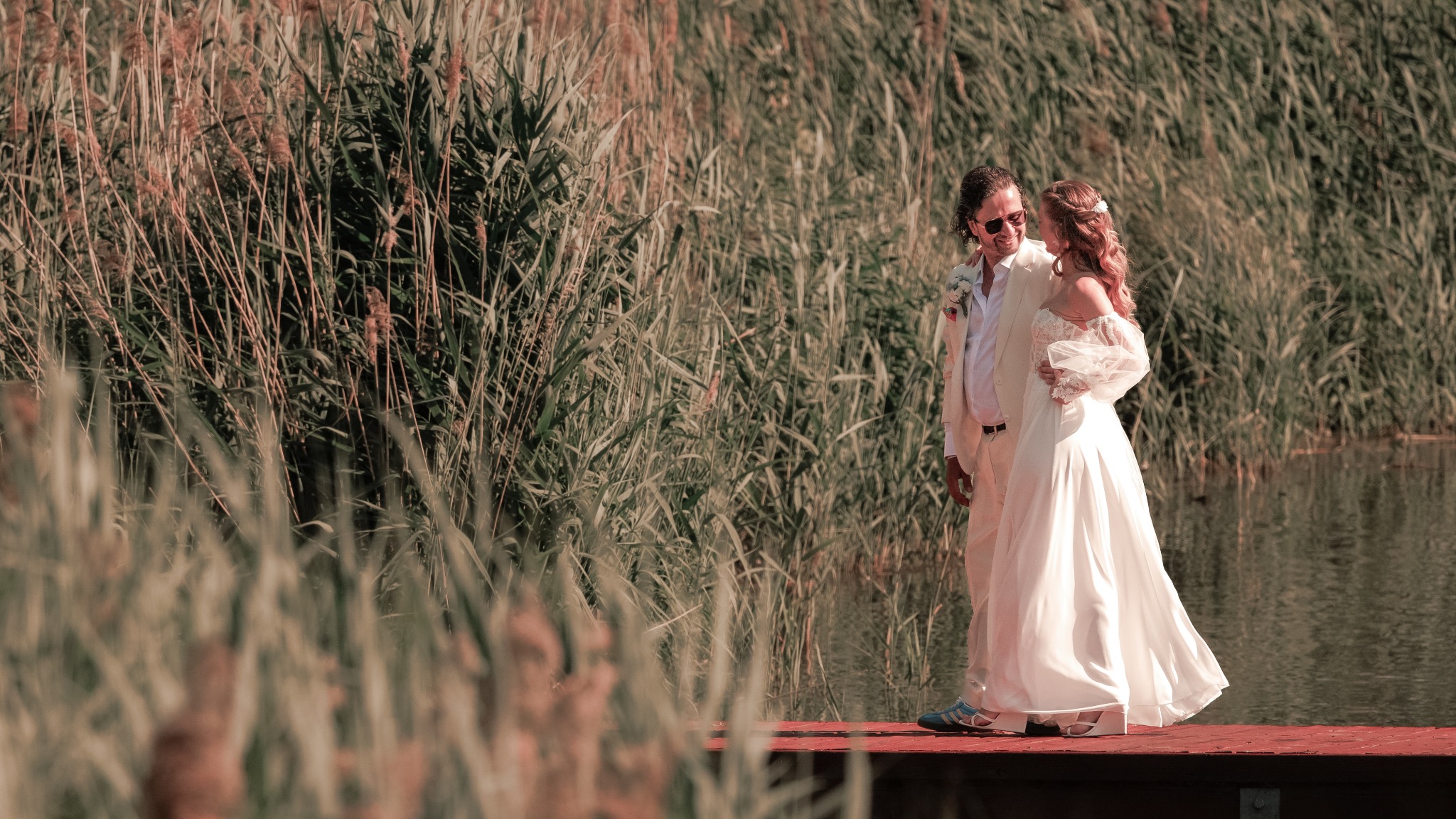 Para młoda w jasnych strojach, stojąca na czerwonym pomoście wśród wysokich traw nad wodą. Pan młody w okularach, panna młoda w sukni z koronkowymi rękawami.