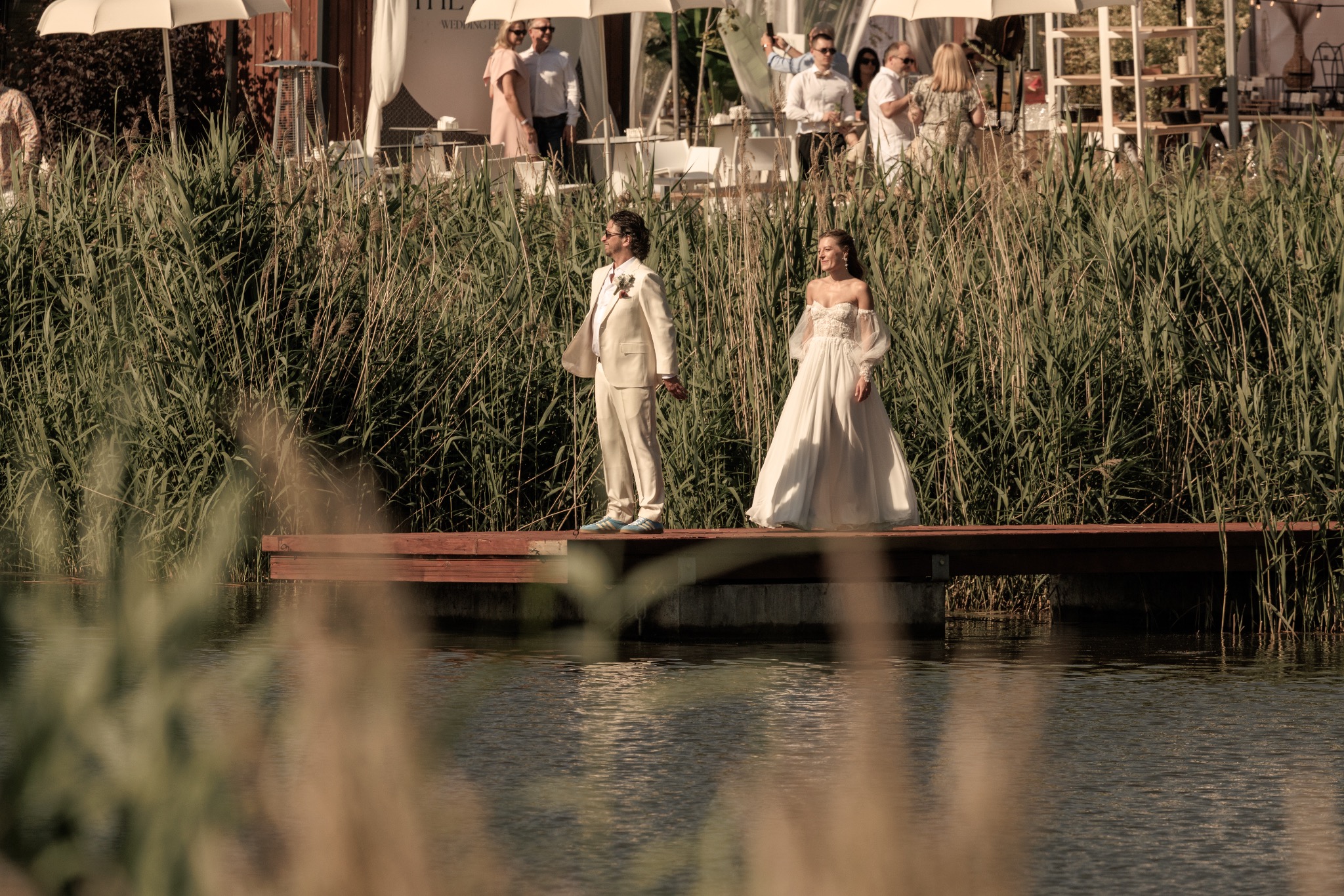 Elegancka para młoda w jasnych strojach pozuje na drewnianym pomoście wśród wysokich traw, z gośćmi weselnymi w tle. Ujęcie w naturalnym świetle.