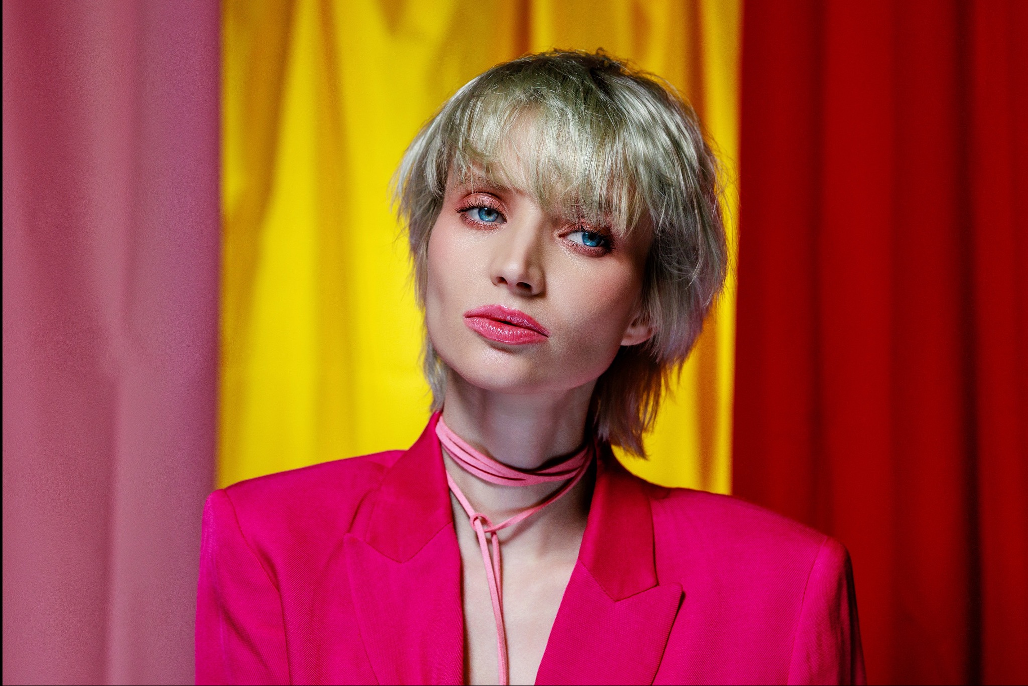Portret kobiety w różowym żakiecie na tle kolorowych zasłon. Krótkie blond włosy, wyrazisty makijaż oczu i ust. Profesjonalna sesja w studio.