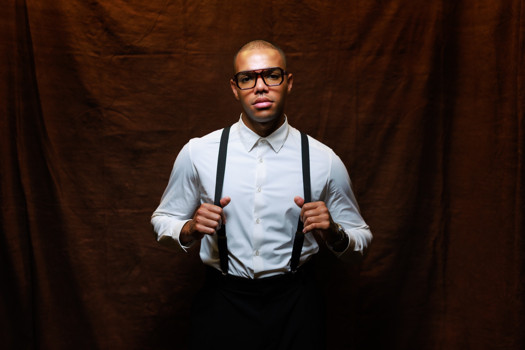 Mężczyzna w okularach i szelkach na tle brązowej tkaniny. Portret w studiu, oświetlenie podkreśla rysy twarzy i fakturę materiału.