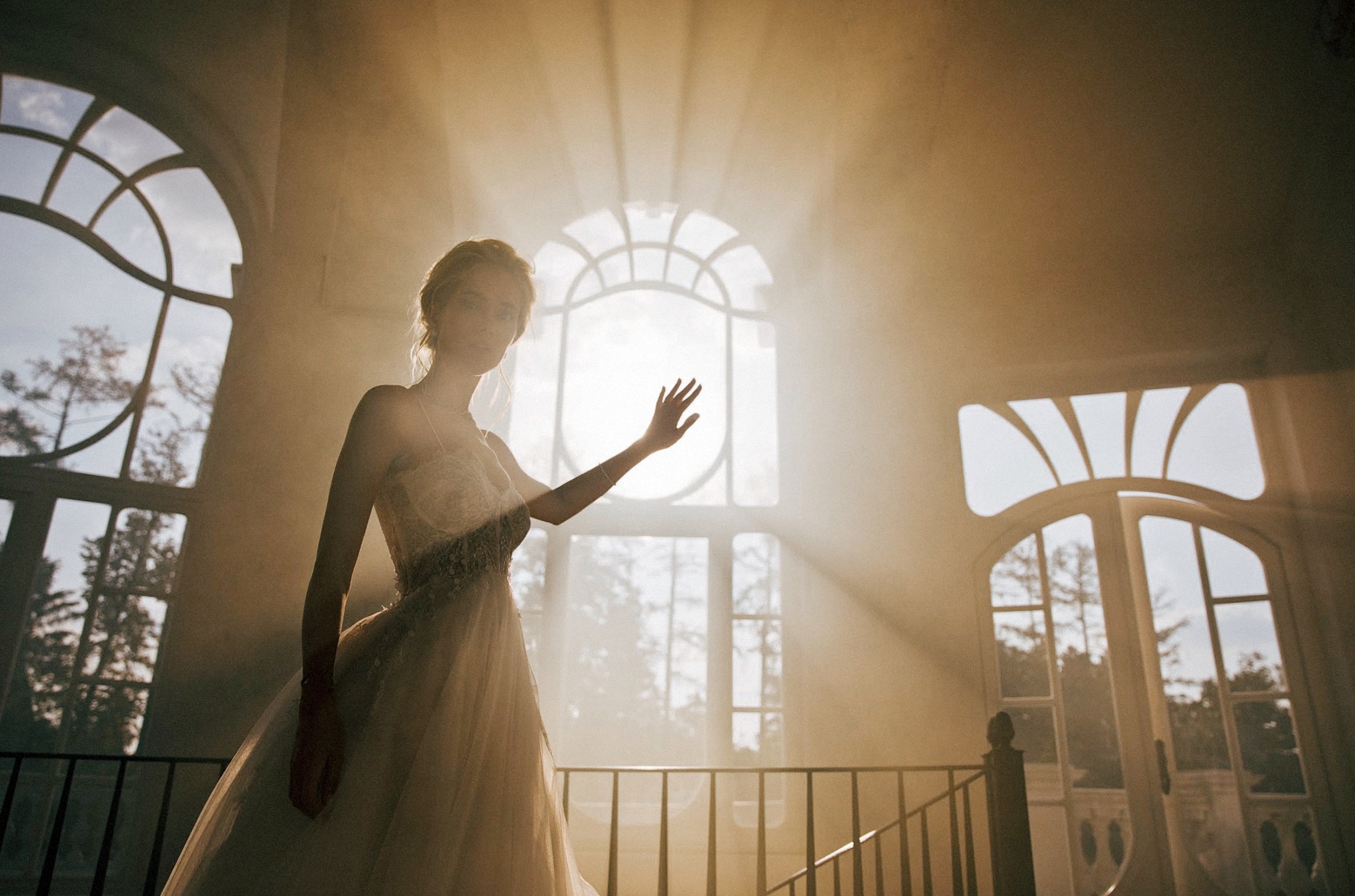 Panna młoda w sukni ślubnej, oświetlona promieniami słońca wpadającymi przez okna o łukowatych ramach. Delikatna sylwetka na tle jasnego wnętrza.