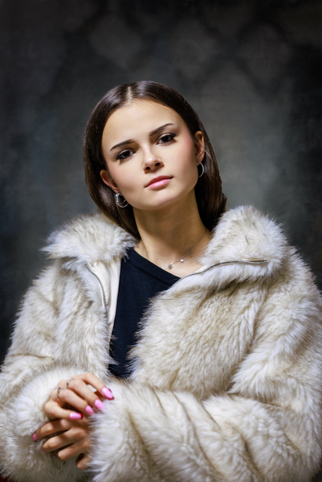 Portret młodej kobiety w futrze na ciemnym tle, z delikatnym makijażem i biżuterią, dłonie splecione. Ujęcie od dołu podkreśla elegancję i styl.