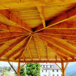 REMONT Z PASJĄ SZYMON KRZUS - Drewniana konstrukcja dachu altany ogrodowej z widocznymi belkami i deskami, w tle budynki i zieleń.