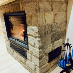 REMONT Z PASJĄ SZYMON KRZUS - Obudowa kominka z kamienia naturalnego z czarną ramą paleniska, widoczne akcesoria kominkowe i drewniana belka stropowa.