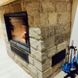 Obudowa kominka z kamienia naturalnego z czarną ramą paleniska, widoczne akcesoria kominkowe i drewniana belka stropowa.