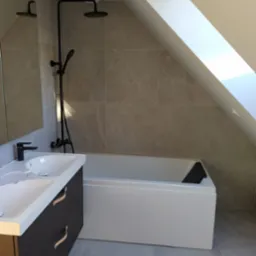 Nowoczesna łazienka na poddaszu z białą wanną, umywalką nablatową na ciemnej szafce z jasnymi uchwytami, szara płytka ścienna i podłogowa, czarna armatura i okno dachowe.