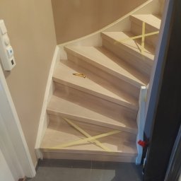 REMONT Z PASJĄ SZYMON KRZUS - Drewniane schody w trakcie renowacji, zabezpieczone taśmą malarską, z widocznym nożykiem introligatorskim na jednym ze stopni.