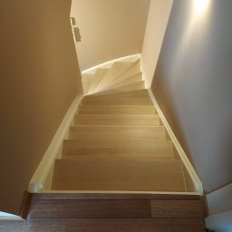 REMONT Z PASJĄ SZYMON KRZUS - Jasne, drewniane schody z białymi listwami przypodłogowymi prowadzące w górę, widok z góry na tle beżowych ścian i podłogi z ciemnego drewna.