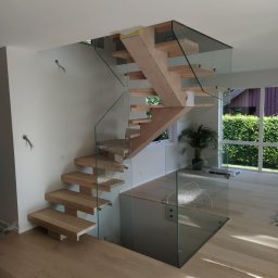 REMONT Z PASJĄ SZYMON KRZUS - Nowoczesne schody drewniane z przezroczystą szklaną balustradą w jasnym wnętrzu domu, widoczne niedokończone detale i wystające przewody elektryczne.