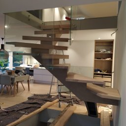 REMONT Z PASJĄ SZYMON KRZUS - Montaż drewnianych schodów z szklaną balustradą w nowoczesnym wnętrzu, widoczne narzędzia i laserowe wyznaczniki poziomu.