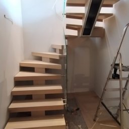 REMONT Z PASJĄ SZYMON KRZUS - Nowoczesne schody drewniane z szklaną balustradą w trakcie montażu, widoczna drabina i narzędzia na podłodze.
