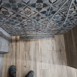 REMONT Z PASJĄ SZYMON KRZUS - Ukończona kabina prysznicowa z odpływem liniowym, wykończona płytkami imitującymi drewno na podłodze i wzorzystymi płytkami na ścianach, widoczne buty robocze w dolnej części kadru.