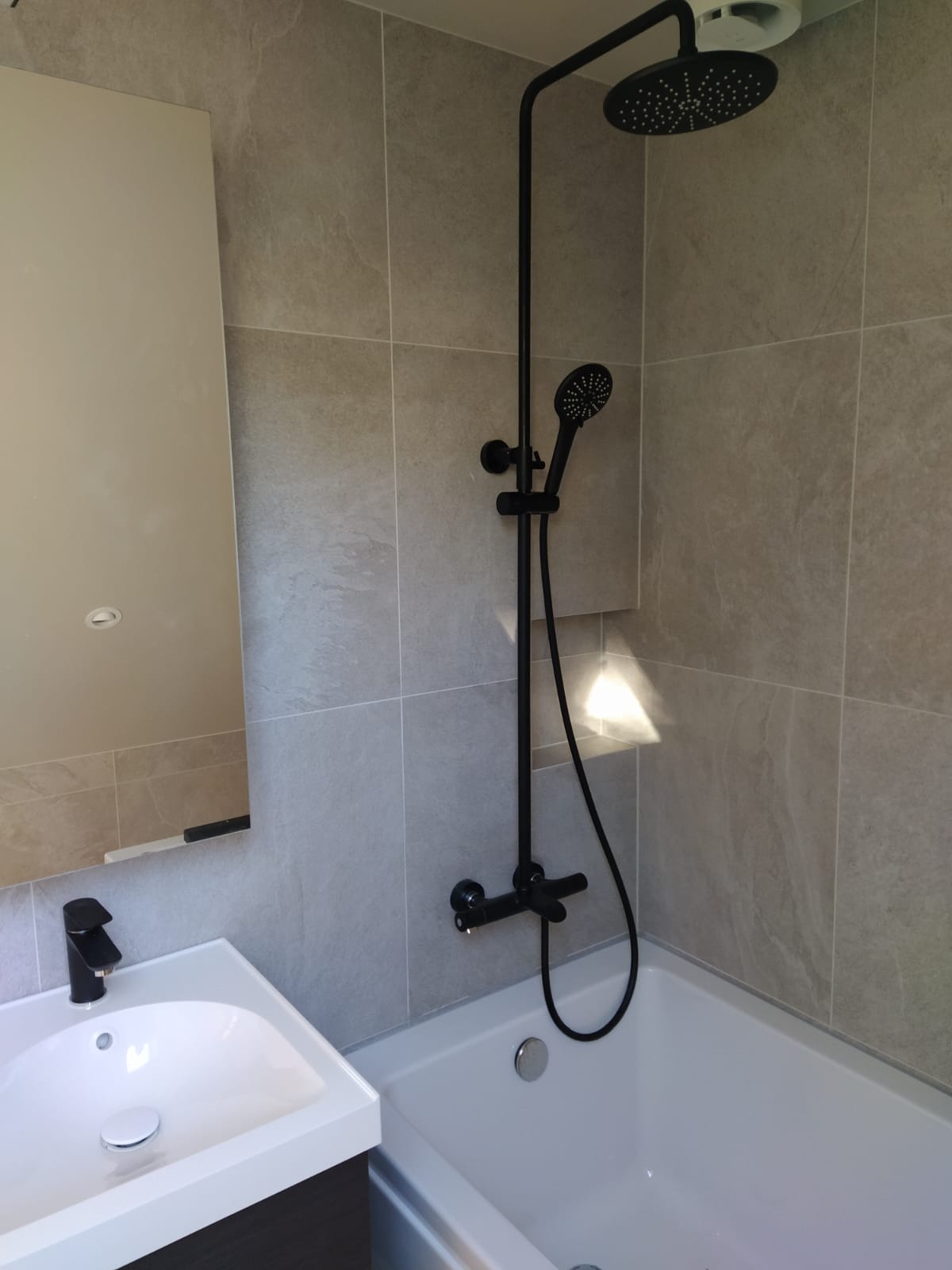 Nowoczesna łazienka z szarymi płytkami, czarną armaturą i białą wanną oraz umywalką z szafką, oświetlona światłem padającym z góry.