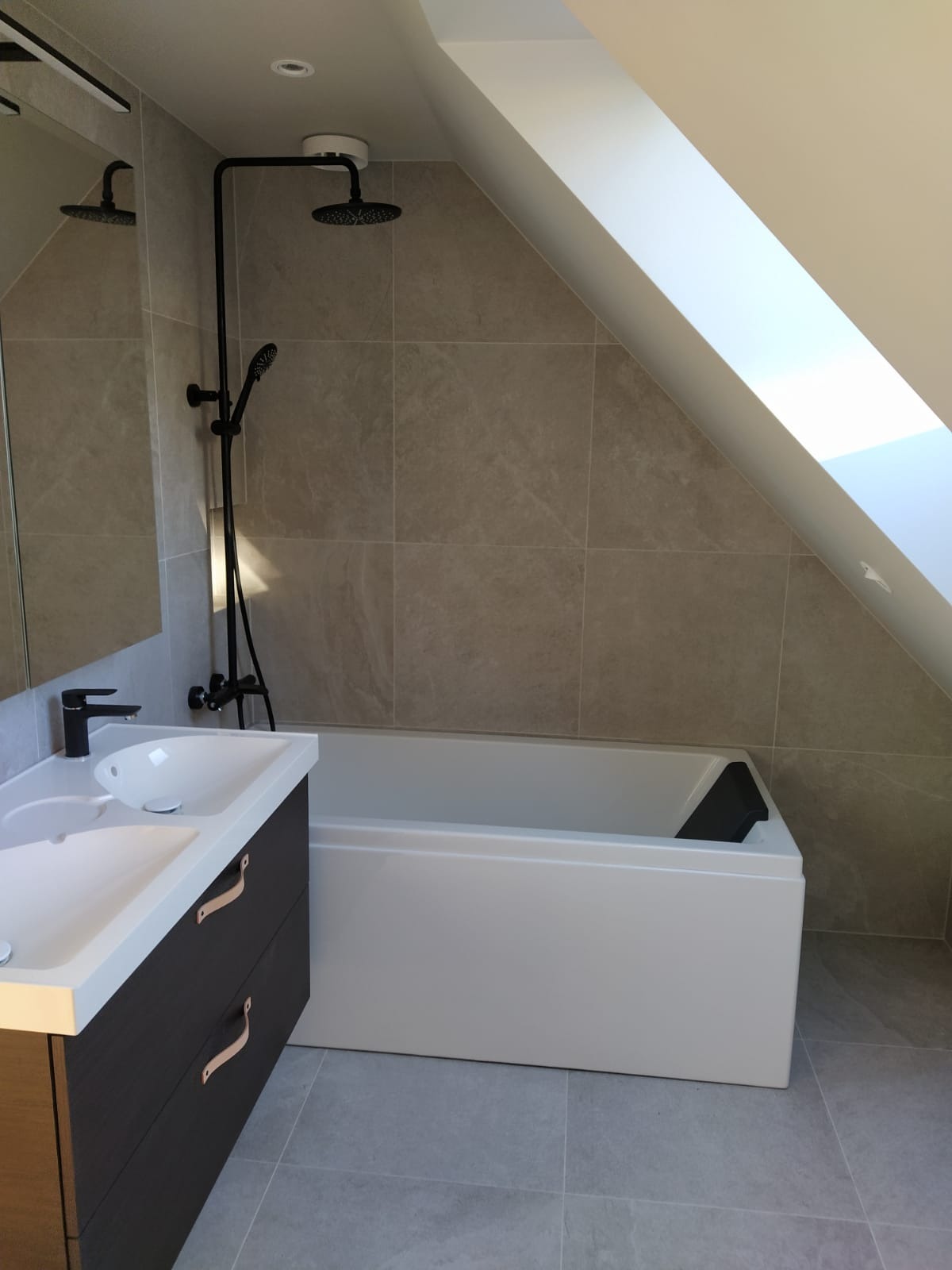 Nowoczesna łazienka na poddaszu z białą wanną, umywalką nablatową na ciemnej szafce z jasnymi uchwytami, szara płytka ścienna i podłogowa, czarna armatura i okno dachowe.