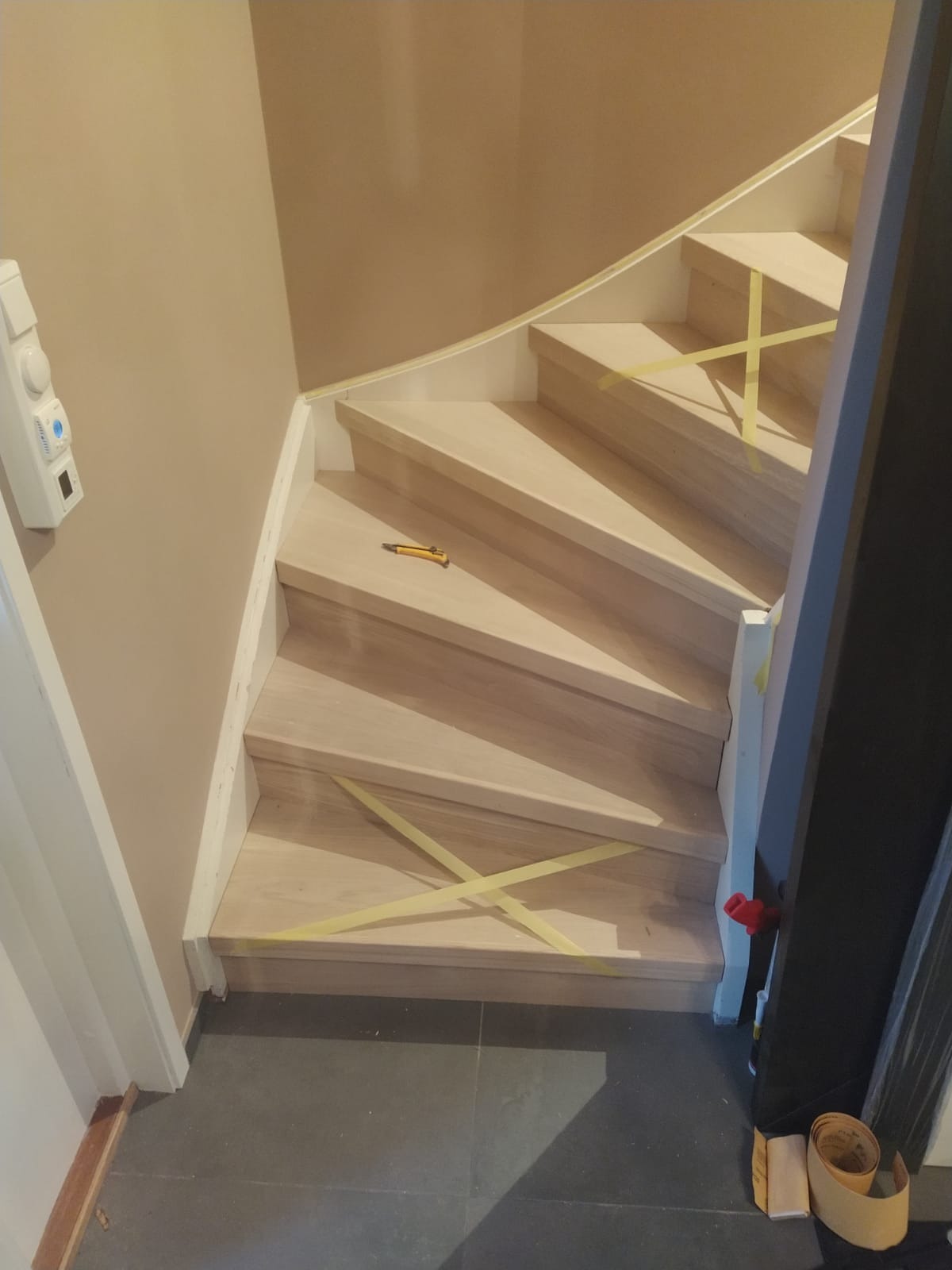 Drewniane schody w trakcie renowacji, zabezpieczone taśmą malarską, z widocznym nożykiem introligatorskim na jednym ze stopni.
